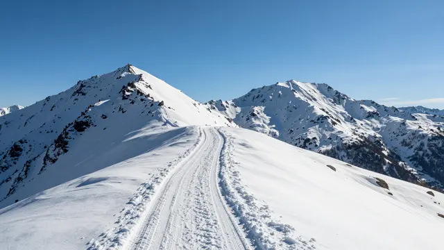 A groomed snowy mountain ridge trail leads toward two distinct snow-covered peaks under a bright clear blue sky