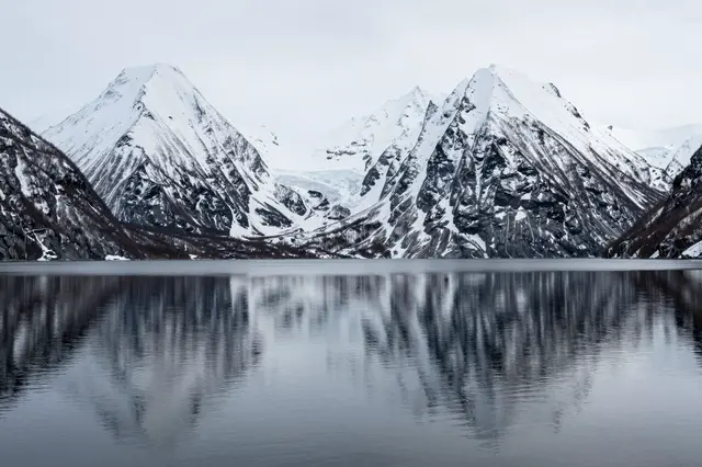 Sharp snow-capped mountain range reflects perfectly in a still, quiet alpine lake under an overcast sky