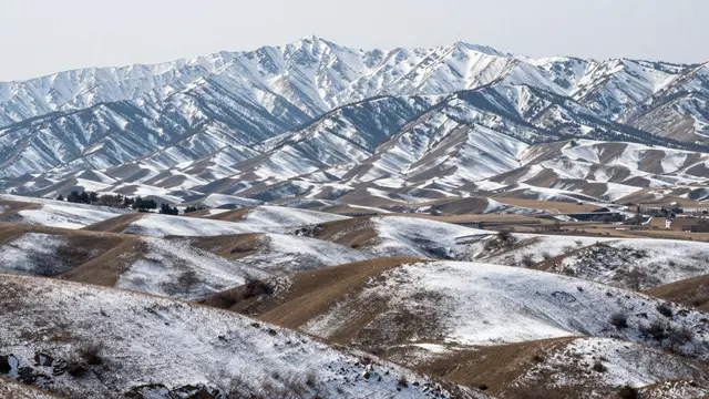 Patchy snow covers rolling grassy foothills leading up to rugged snow-capped mountains under an overcast sky