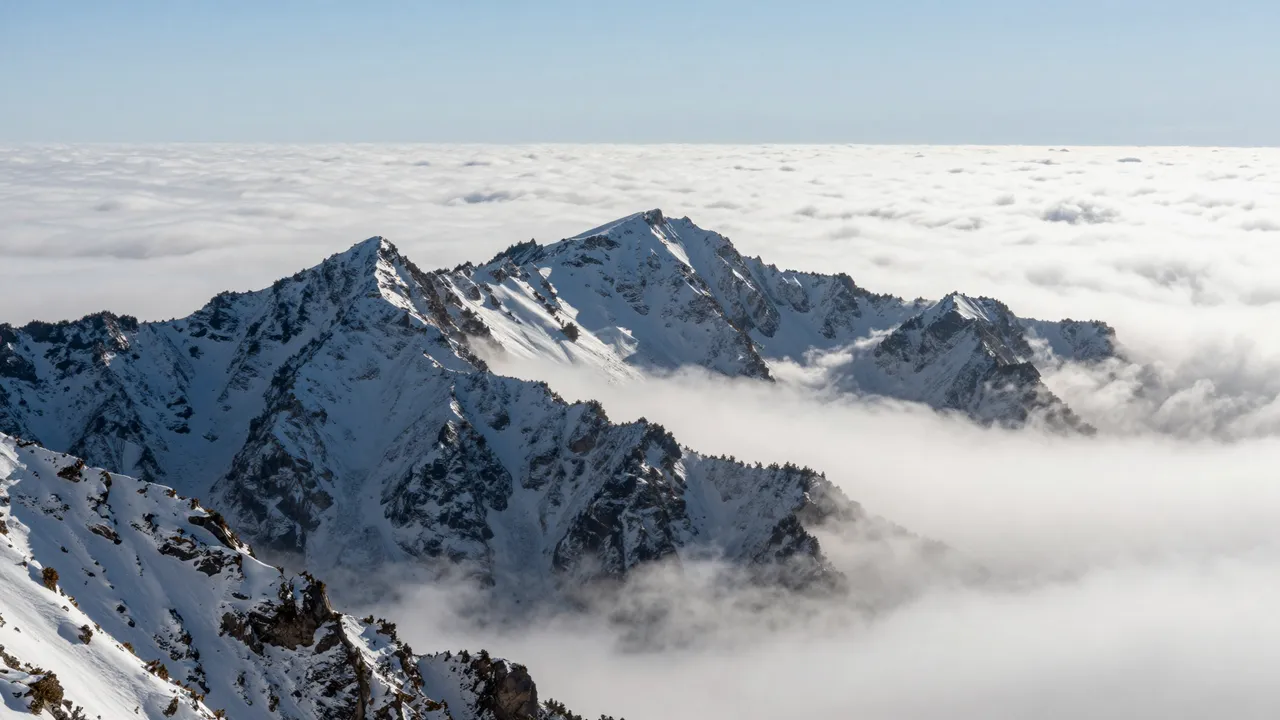 Rugged snow covered mountain peaks rise above a thick sea of white clouds under a pale clear blue sky