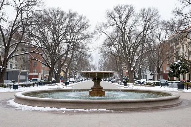 A circular stone fountain sits in a snow-dusted paved urban plaza lined with bare winter trees