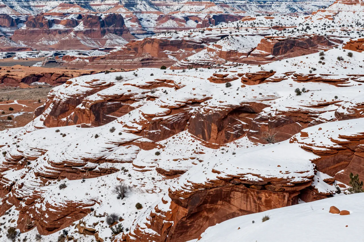 Layered red desert canyon rock formations dusted with fresh winter snow stretch across the landscape