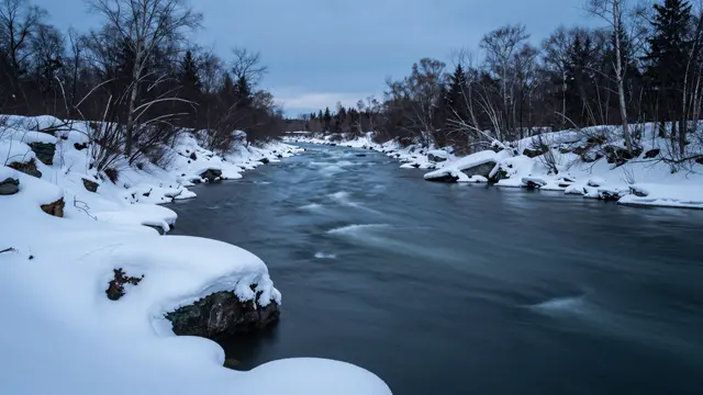 Long exposure view of a dark flowing river flanked by snow-covered banks during cold blue hour twilight