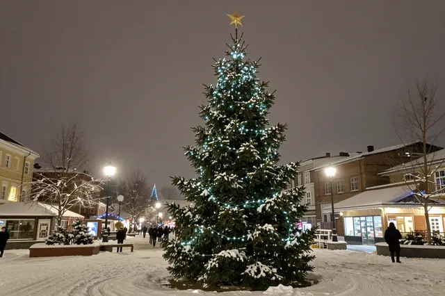 A lit snow-dusted Christmas tree stands in a snowy town square on a dark winter night