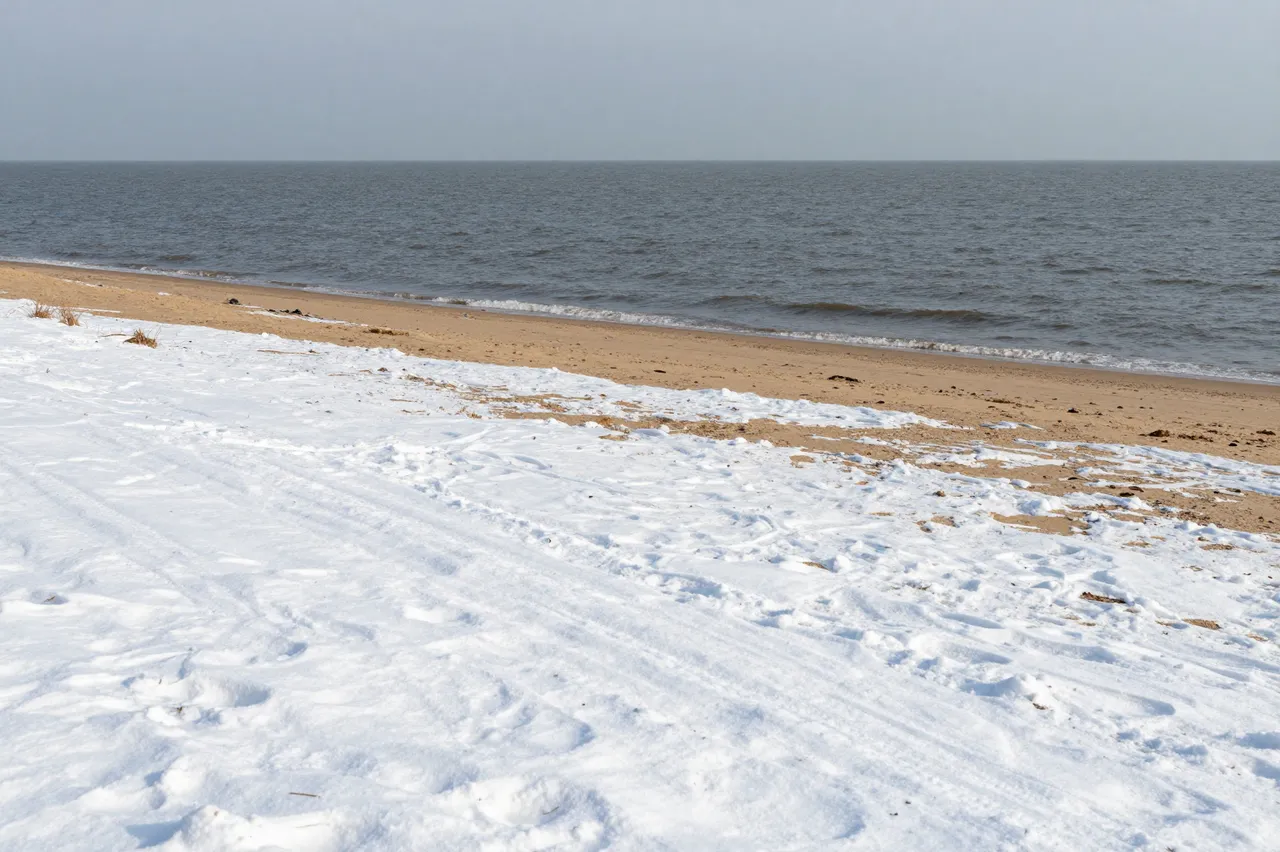 Patchy snow covers a sandy beach beside a calm gray sea under an overcast sky