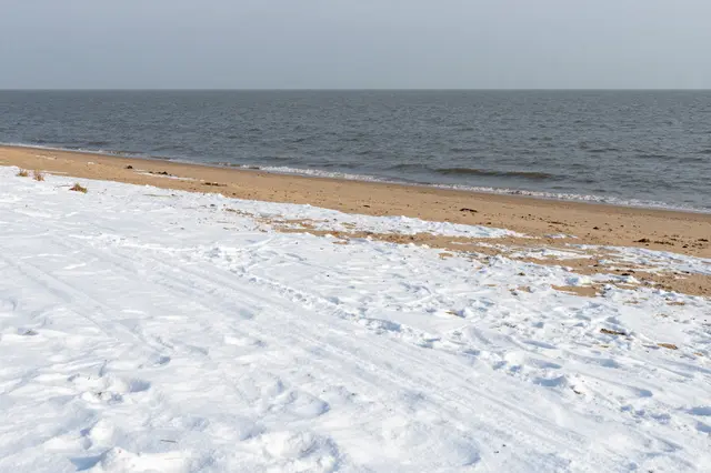 Patchy snow covers a sandy beach beside a calm gray sea under an overcast sky
