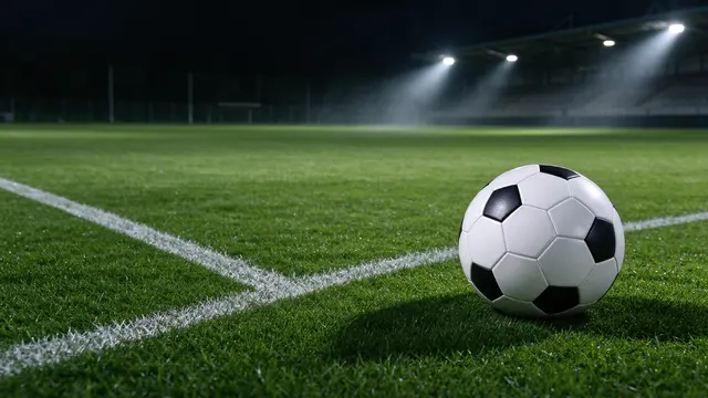 A classic black and white soccer ball rests on floodlit grass at a football pitch corner at night.