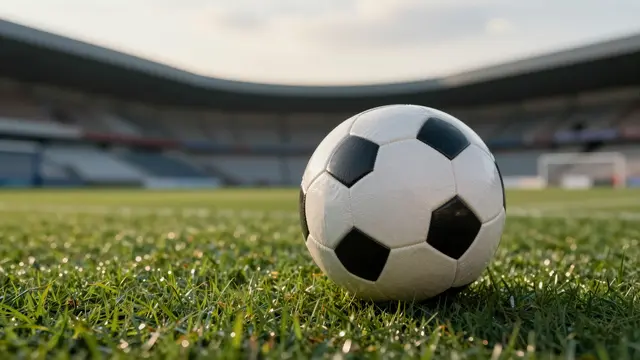 A classic black and white soccer ball rests on dewy green grass in an empty soccer stadium at soft sunset.
