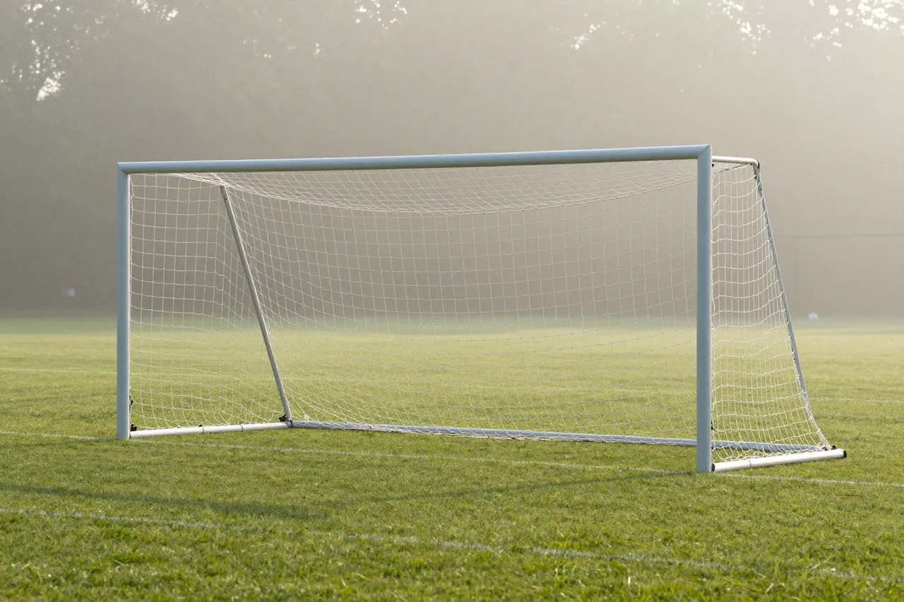 A soccer goal net sits on an empty grass field surrounded by soft morning fog