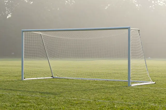 A soccer goal net sits on an empty grass field surrounded by soft morning fog