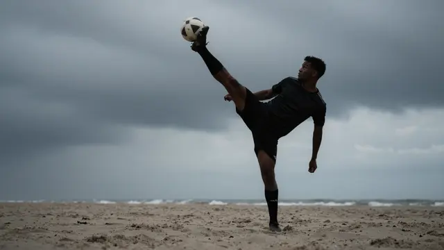 Adult male soccer player balances a soccer ball on his raised foot on a sandy beach under a stormy gray sky