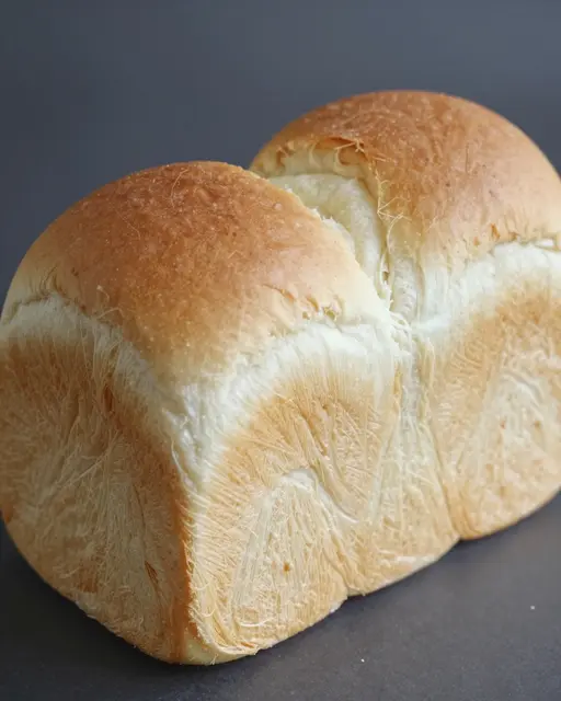 A plump golden crust Japanese milk bread loaf rests on a dark matte surface.