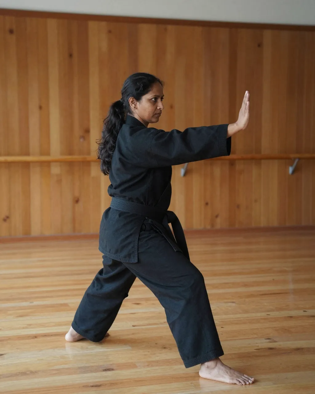 Side view of a South Asian woman in a black karate uniform performing a stance in a wooden dojo