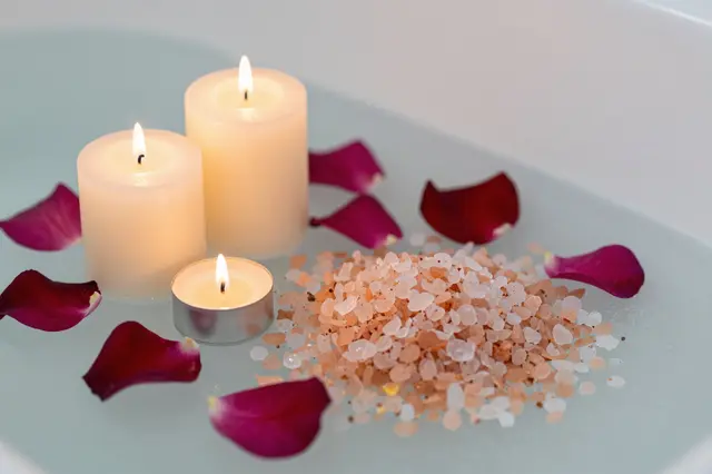 Lit candles, pink bath salts, and rose petals rest on the surface of a filled bathtub