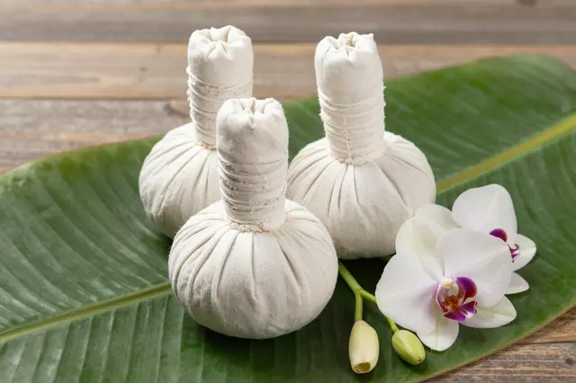 Three herbal massage compress balls rest on a banana leaf with white orchids over a wooden table.
