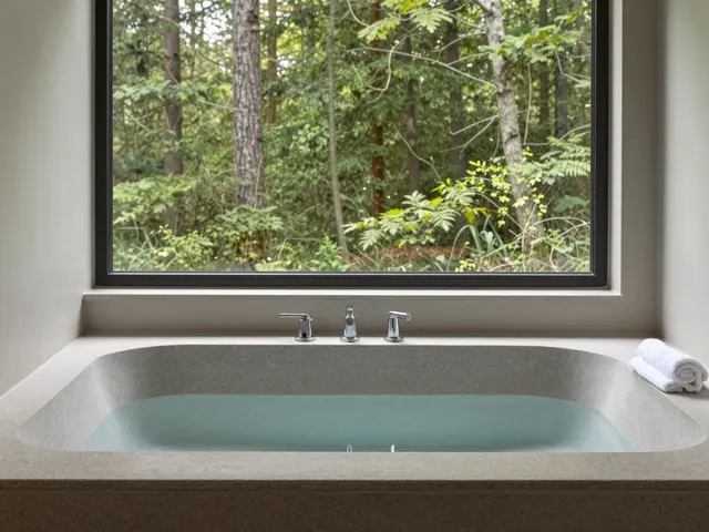 A filled speckled stone bathtub sits below a large window overlooking a lush green forest.