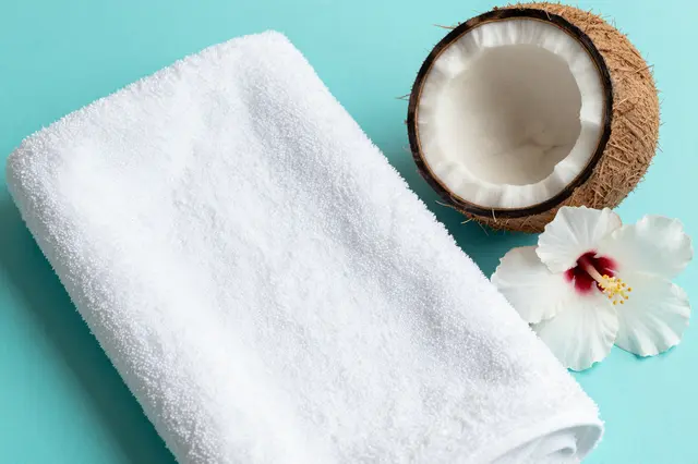 Top view of a folded white towel, halved coconut, and white hibiscus flower on a turquoise solid surface