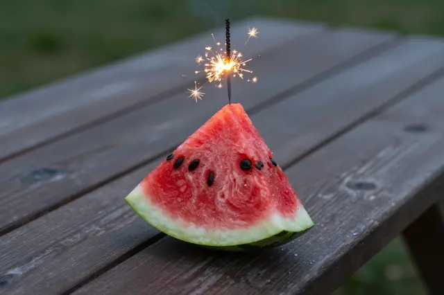 A lit sparkler is stuck in a watermelon wedge resting on a wooden picnic table at dusk