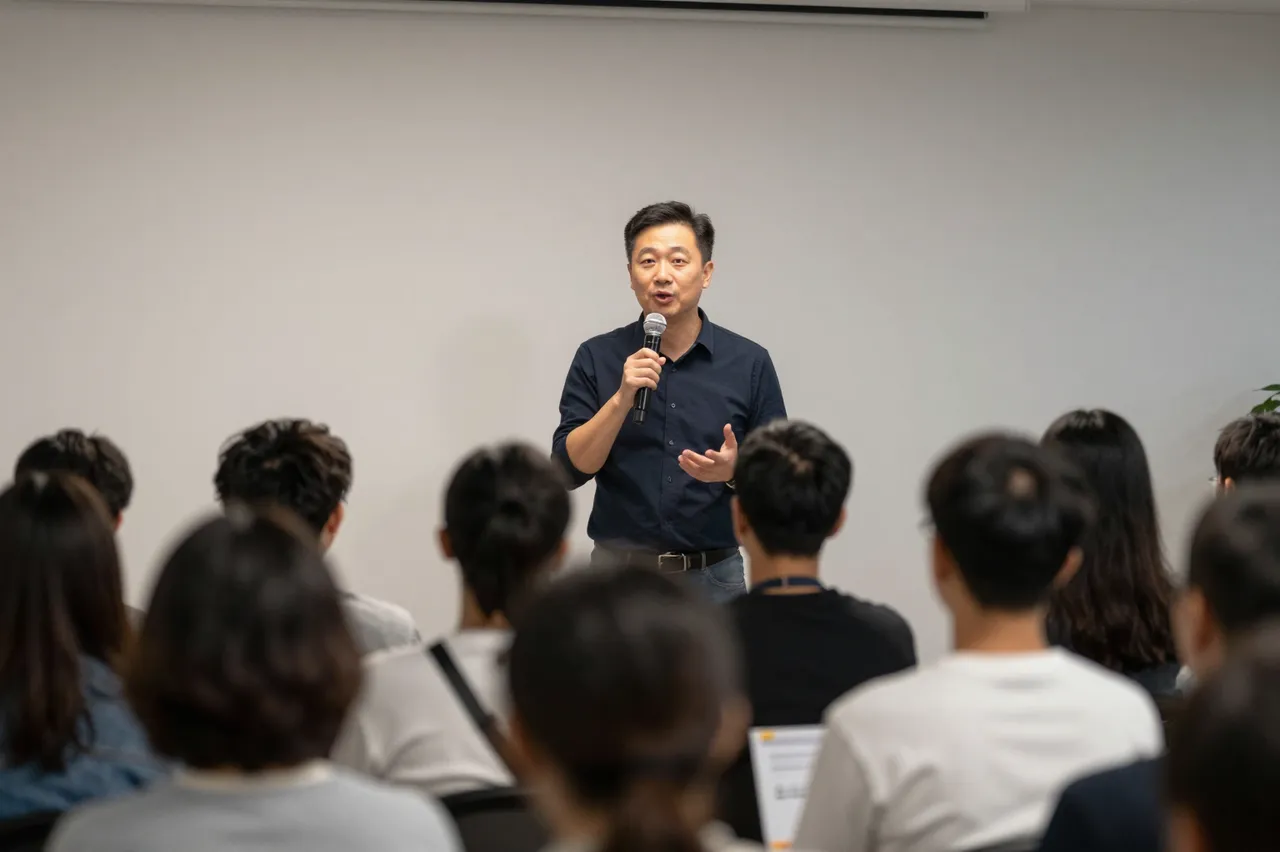 A male speaker holding a microphone addresses a seated audience viewed from behind in an indoor meeting room