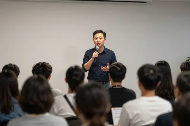 A male speaker holding a microphone addresses a seated audience viewed from behind in an indoor meeting room