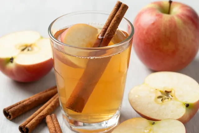 Glass of apple cider with cinnamon sticks and fresh sliced apples arranged on a light tabletop