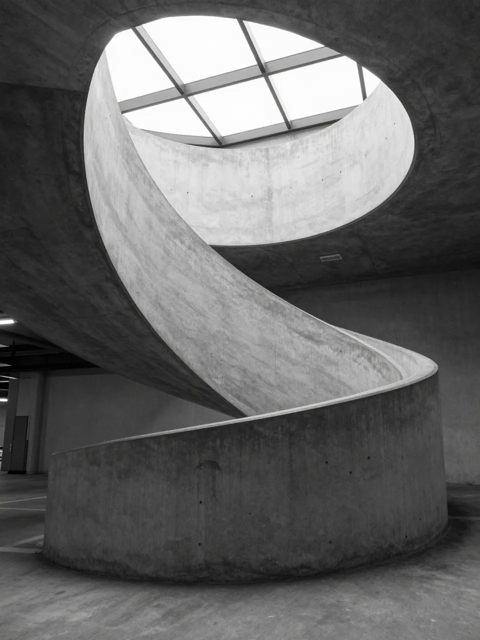 Black and white view of a curved concrete spiral ramp rising toward a circular skylight inside an empty parking garage