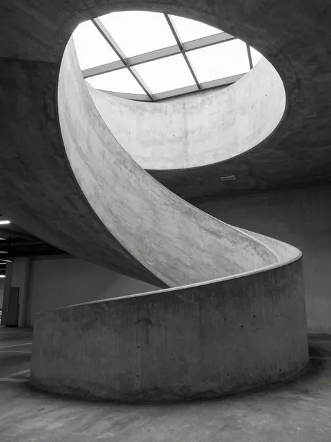 Black and white view of a curved concrete spiral ramp rising toward a circular skylight inside an empty parking garage