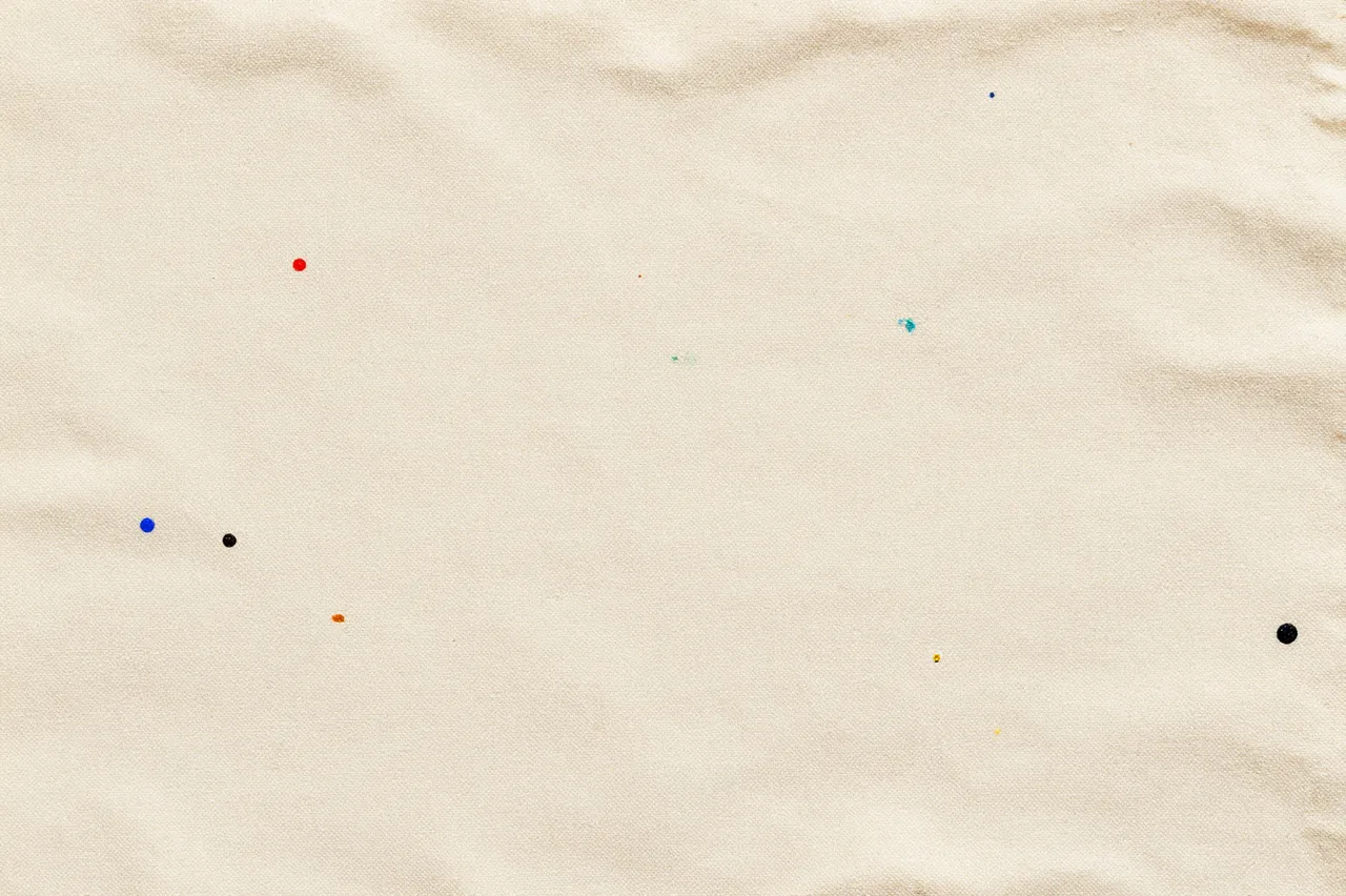 Top view of a textured beige cotton drop cloth scattered with small multicolored dried paint splatters.