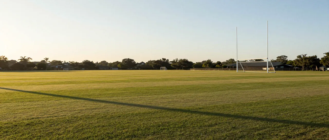 Empty grass sports field with goal posts under warm golden hour sunlight stretching long shadows across the turf