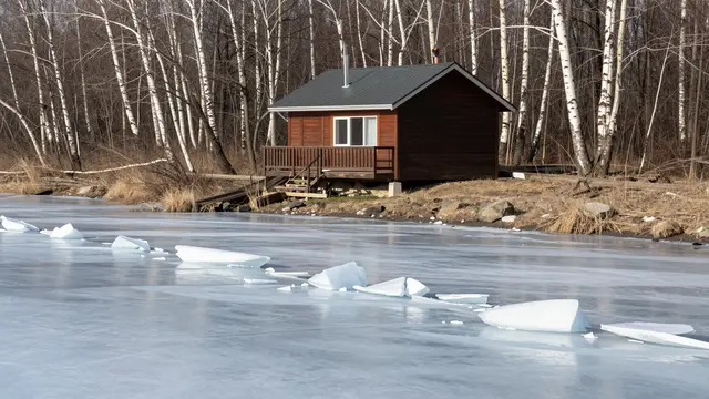 Small wooden cabin sits on a lakeshore beside broken ice chunks and bare birch trees during spring thaw