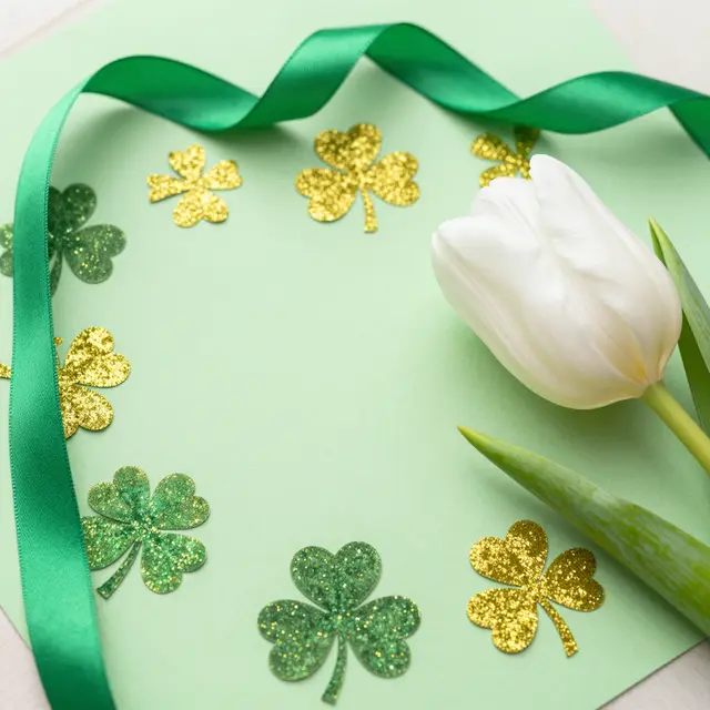 St. Patrick's Day flat lay arrangement with glitter shamrocks, green ribbon and a white tulip on pale green paper
