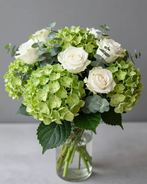Bouquet of green hydrangeas, white roses and eucalyptus arranged in a clear glass vase against a gray background