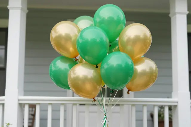 A cluster of shiny green and gold balloons positioned in front of a residential house porch railing