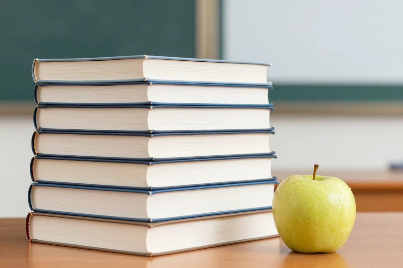 A neat stack of hardcover books sits next to a green apple on a wooden classroom desk