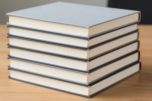 A neat stack of plain hardcover books rests on a light wooden desk surface.