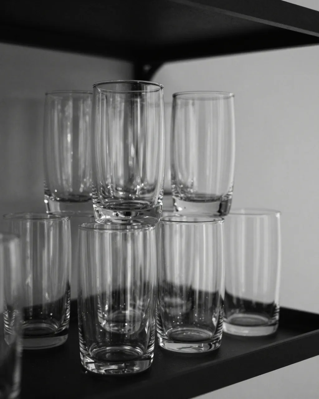 Black and white still life of clear drinking glasses arranged on a dark shelf with light catching their glossy edges.