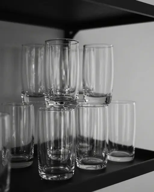 Black and white still life of clear drinking glasses arranged on a dark shelf with light catching their glossy edges.