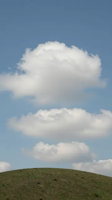 Vertical view of neatly stacked fluffy white clouds above a rounded green hill under clear blue sky