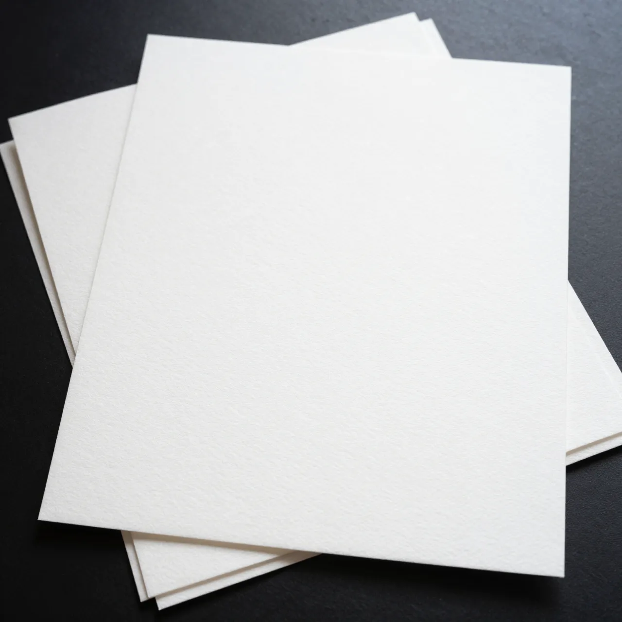 Overlapping sheets of bright white hot-press watercolor paper rest on a dark smooth surface