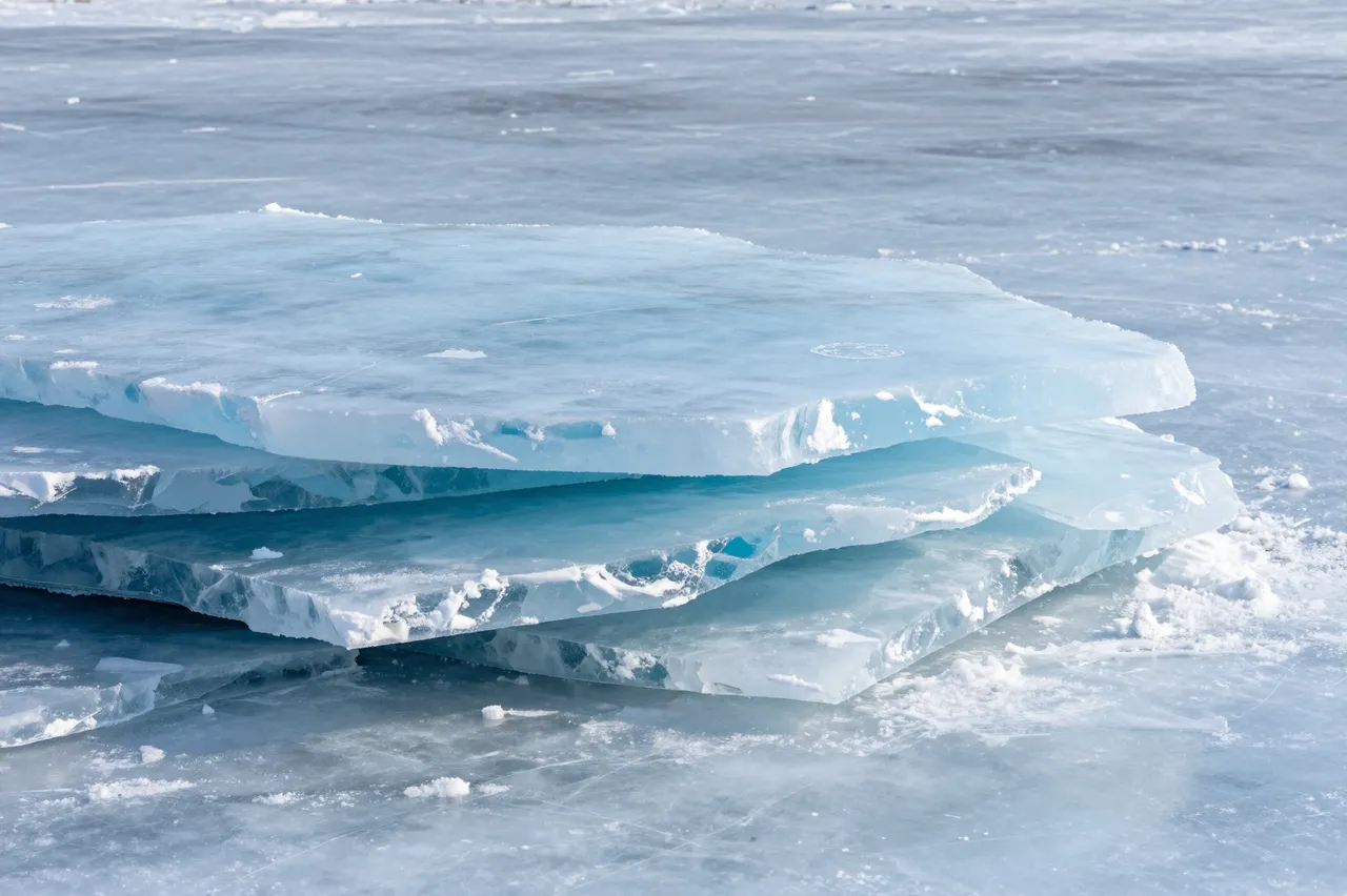 Piled clear blue ice slabs rest on the smooth surface of a frozen lake under bright daylight