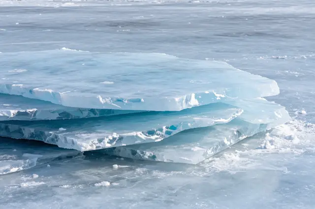 Piled clear blue ice slabs rest on the smooth surface of a frozen lake under bright daylight