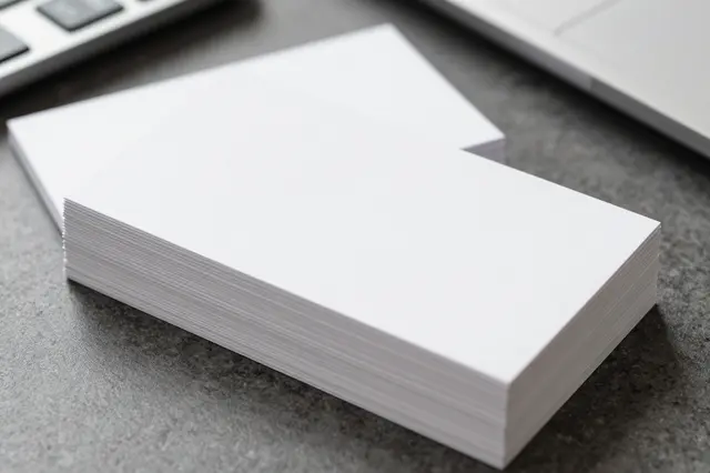 Two stacks of blank white business cards sit on a dark office desk near a keyboard and laptop edge.