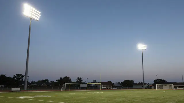 Tall glowing stadium floodlights stand over a quiet small-town football and soccer field at twilight