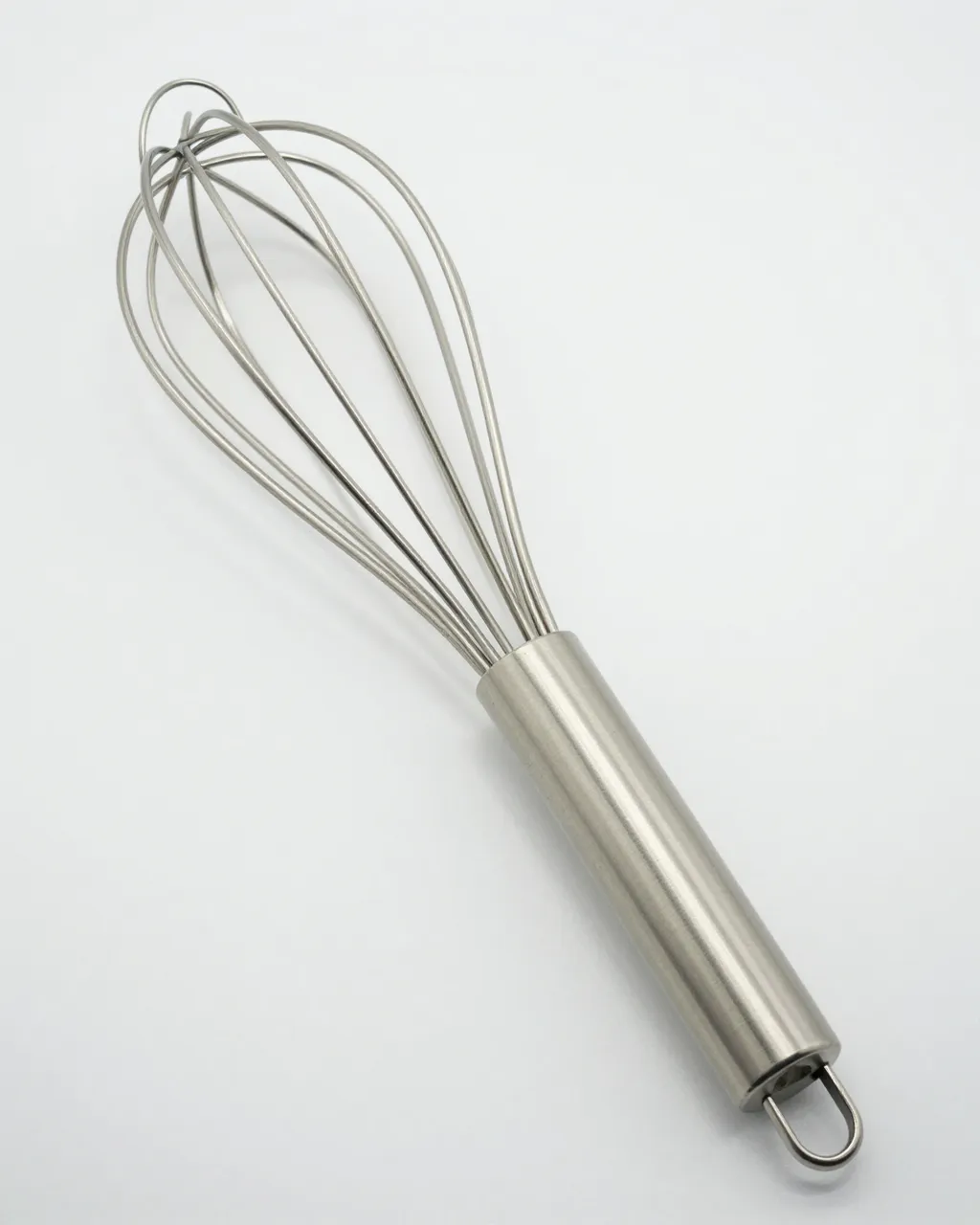 A stainless steel balloon whisk rests at an angle on a plain white studio background.
