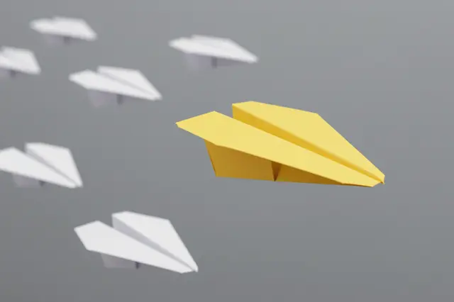A sharp yellow paper plane moves away from a group of blurred white paper planes on a plain gray background