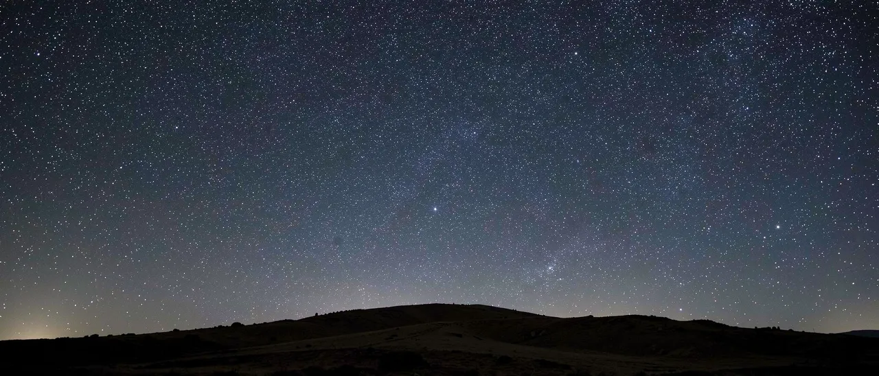 Dense starry night sky stretches above a dark silhouetted low hill ridge with empty upper copy space