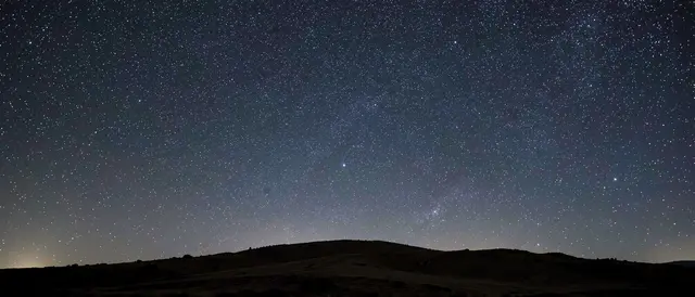 Dense starry night sky stretches above a dark silhouetted low hill ridge with empty upper copy space