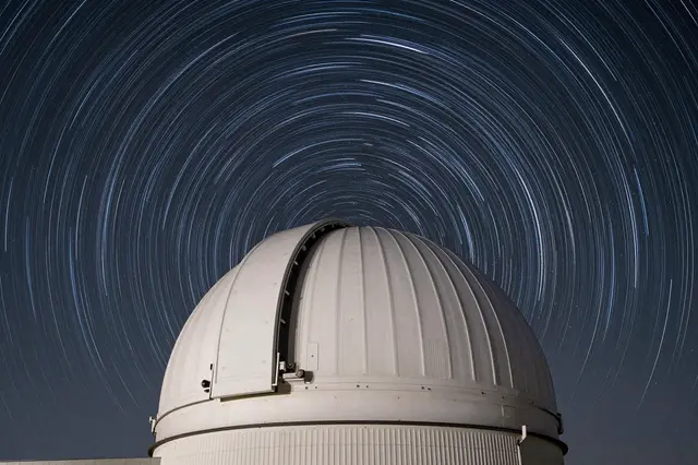 Long exposure circular star trails arc across a dark night sky above a white observatory dome.