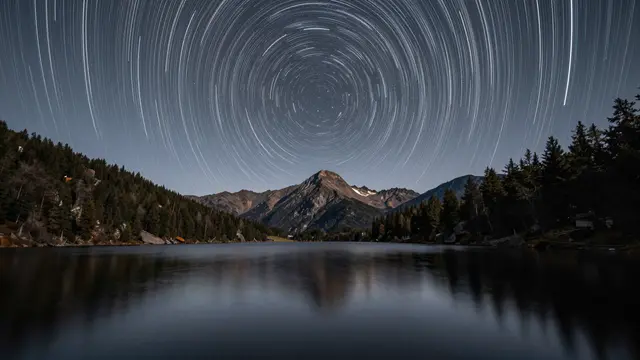 Circular star trails arc across the night sky above a calm mountain lake and forested shorelines