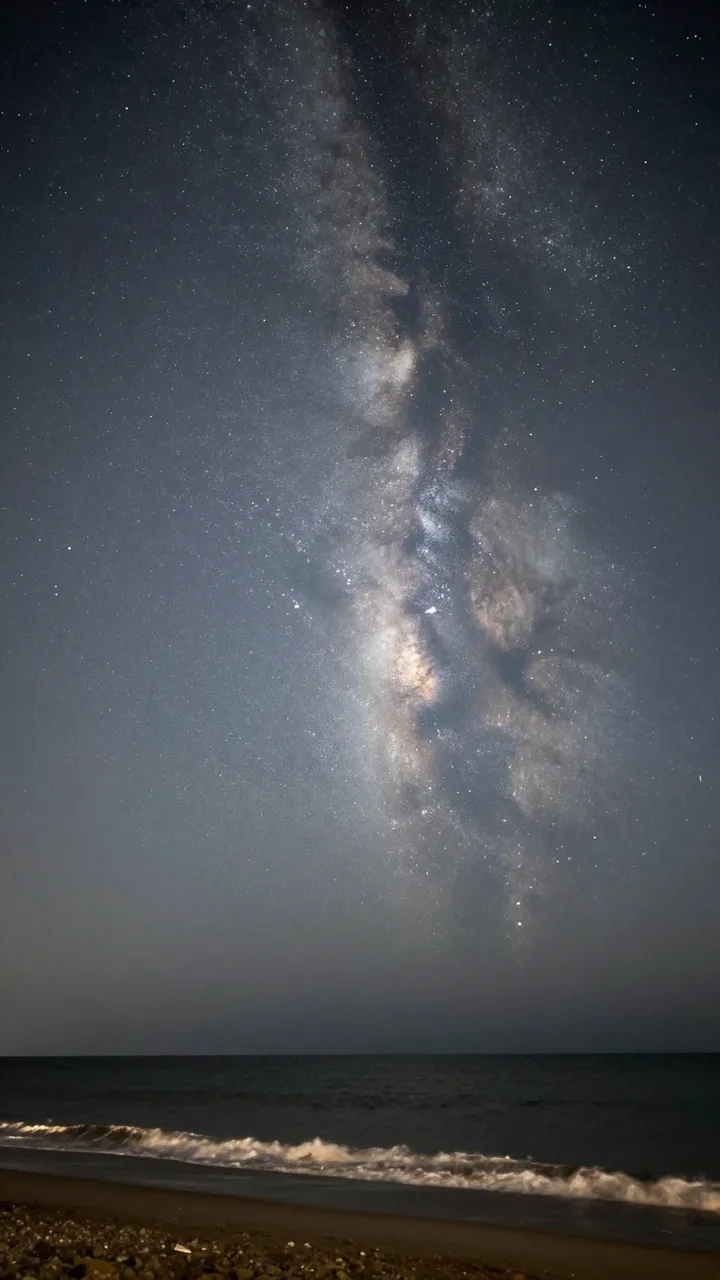 The glowing Milky Way galaxy stretches across a dark night sky above gentle surf on a quiet pebbled beach.